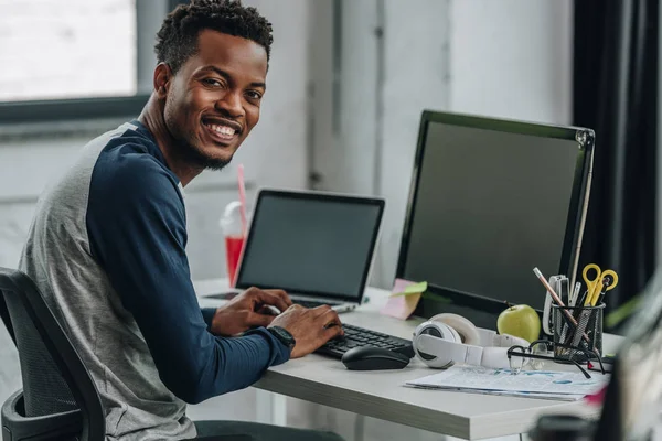 Programmeur afro-américain joyeux regardant la caméra tout en étant assis près des ordinateurs — Photo de stock