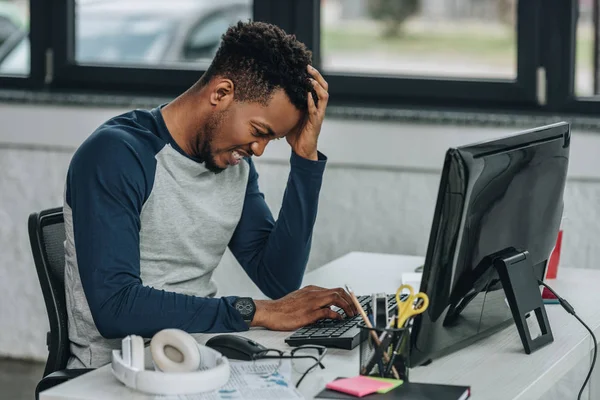 Bouleversé programmeur afro-américain tenant la main près de la tête tout en travaillant sur ordinateur au bureau — Photo de stock