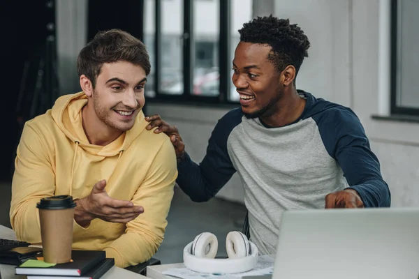 Jovem programador feliz apontando para monitor de computador enquanto sentado perto de colega afro-americano no escritório — Fotografia de Stock