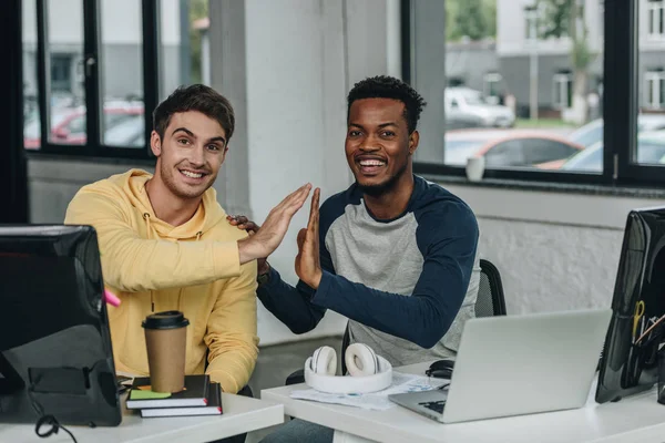 Dos programadores multiculturales dando cinco altos y sonriendo a la cámara en la oficina - foto de stock