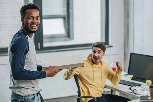 Allegro programmatore afroamericano che tiene la scatola della pizza mentre il collega mostra sì gesto — Foto stock