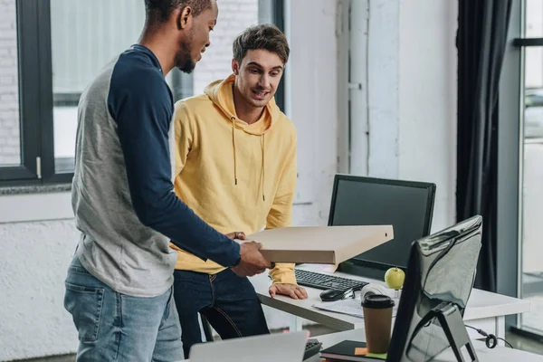 Joven programador afroamericano sosteniendo caja de pizza mientras está de pie cerca de colega sonriente - foto de stock