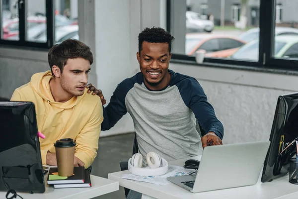 Programador afro-americano alegre apontando para laptop enquanto sentado perto colega — Fotografia de Stock