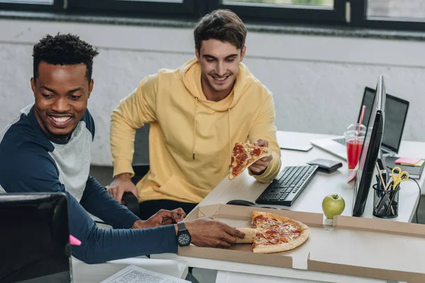 Zwei fröhliche multikulturelle Programmierer, die Pizza essen, während sie im Büro am Schreibtisch sitzen — Stockfoto