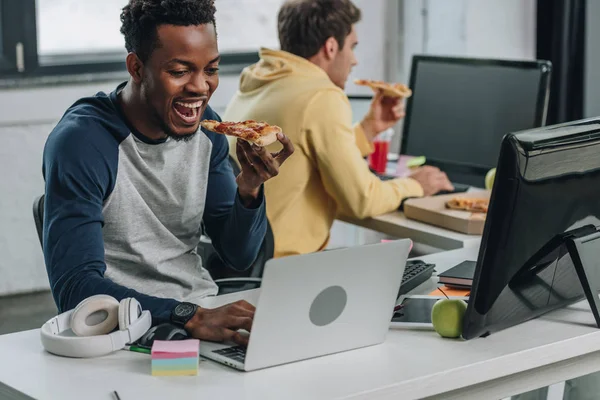 Zwei multikulturelle Programmierer, die Pizza essen, während sie im Büro neben Computern sitzen — Stockfoto