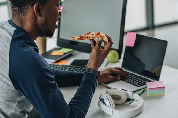 Afrikanischer amerikanischer Programmierer isst Pizza während er am Laptop arbeitet — Stockfoto