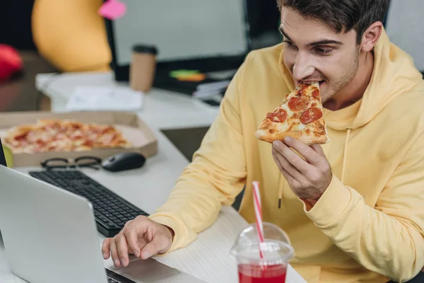 Bello programmatore mangiare pizza mentre seduto sul posto di lavoro in ufficio — Foto stock