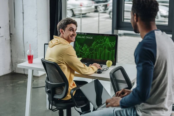 Веселий програміст сидить на моніторі комп'ютера з онлайн-торгівлею на екрані і дивиться на афроамериканського колегу — стокове фото