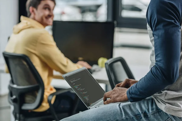 Vista recortada del programador afroamericano trabajando mientras está sentado en el escritorio cerca de un colega sonriente - foto de stock