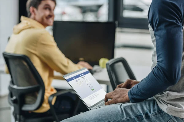 KYIV, UKRAINE - 29 JUILLET 2019 : vue recadrée d'un programmeur afro-américain utilisant un ordinateur portable avec le site Facebook à l'écran près d'un collègue souriant — Photo de stock