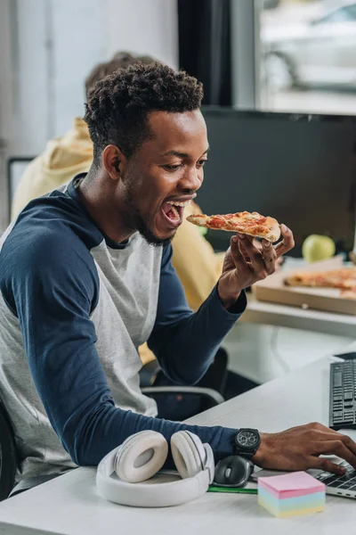 Programador afro-americano faminto comer pizza enquanto sentado no local de trabalho perto colega — Fotografia de Stock