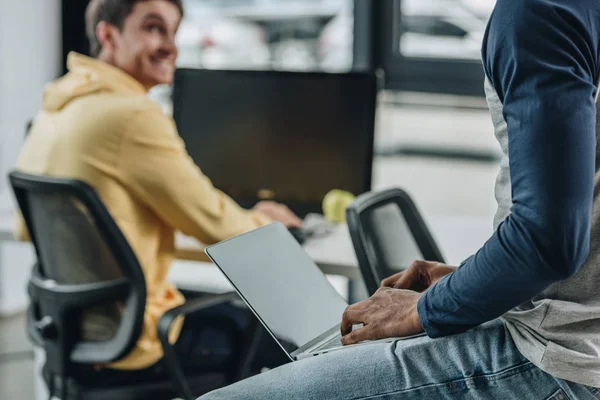 Partial view of african american programmer sitting on desk with laptop near smiling colleague — Stock Photo