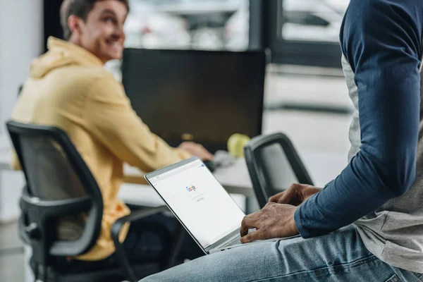 KYIV, UKRAINE - 29 JUILLET 2019 : vue recadrée d'un programmeur afro-américain tenant un ordinateur portable avec Google à l'écran près d'un collègue souriant — Photo de stock