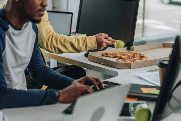 Abgeschnittene Ansicht eines Programmierers, der ein Stück Pizza in der Nähe eines afrikanischen amerikanischen Kollegen hält — Stockfoto