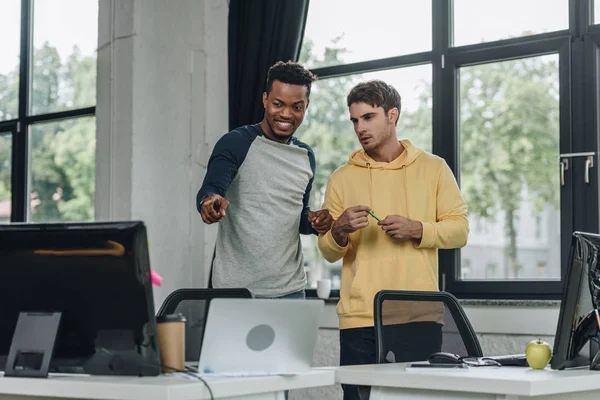 Cheerful african american programmer pointing with finger at computer monitor while standing near colleague — Stock Photo