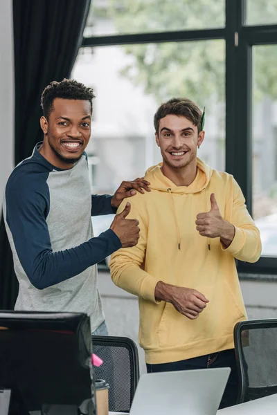 Two happy multicultural programmer showing thumbs up while smiling at camera in office — Stock Photo