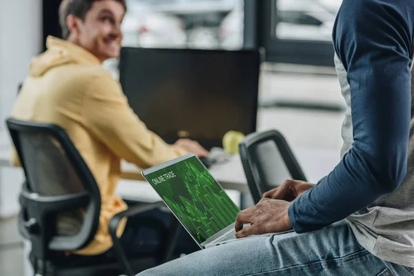 Abgeschnittene Ansicht eines afrikanisch-amerikanischen Programmierers, der am Tisch sitzt und Laptop mit Online-Handel am Bildschirm in der Nähe eines multikulturellen Kollegen hält — Stockfoto