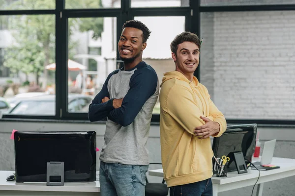 Dos programadores multiculturales felices parados espalda a espalda con los brazos cruzados y sonriendo a la cámara en la oficina - foto de stock