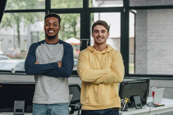 Two cheerful multicultural programmers standing with crossed arms and smiling at camera in office — Stock Photo