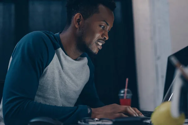 Sonriente programador afroamericano sentado en el lugar de trabajo por la noche - foto de stock