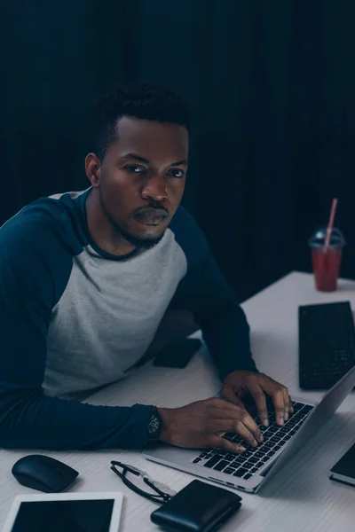 Programmeur afro-américain sérieux regardant la caméra tout en travaillant la nuit au bureau — Photo de stock