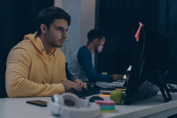 Foco selectivo de programador serio mirando monitor mientras que trabaja en la noche cerca de amigo afroamericano - foto de stock