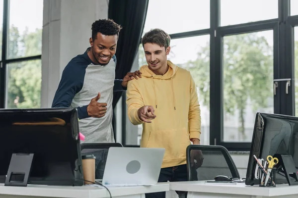 Dos programadores multiculturales alegres mirando el ordenador portátil en la oficina - foto de stock