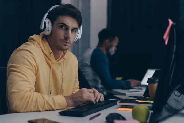Enfoque selectivo de un joven programador en auriculares mirando a la cámara mientras trabaja cerca de un colega afroamericano - foto de stock