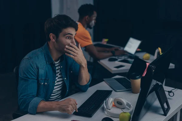 Selective focus of surprised programmer looking at monitor while working near african american colleague — Stock Photo