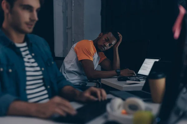 Enfoque selectivo del programador afroamericano mirando a un colega multicultural mientras trabajan juntos por la noche en la oficina - foto de stock