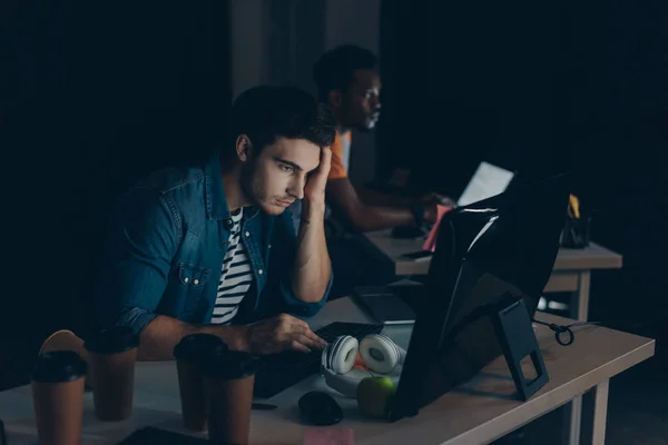 Selektiver Fokus eines durchdachten Programmierers, der nachts im Büro in der Nähe eines afrikanischen amerikanischen Kollegen arbeitet — Stockfoto