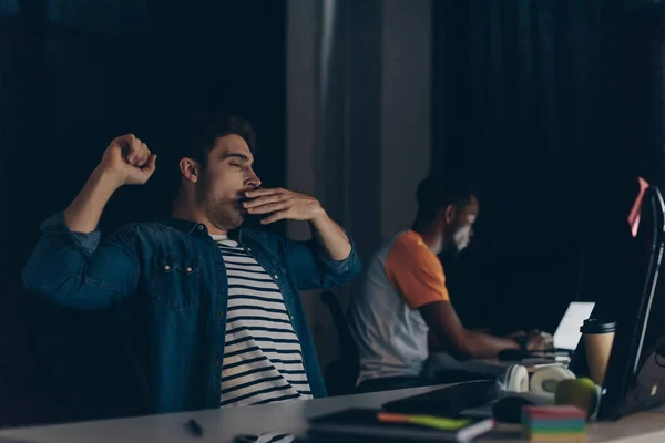 Selective focus of tired programmer yawning while sitting near african american colleague and night in office — Stock Photo