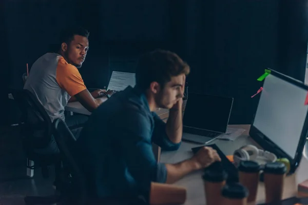 Enfoque selectivo del programador afroamericano mirando a un colega multicultural mientras trabaja de noche en la oficina - foto de stock