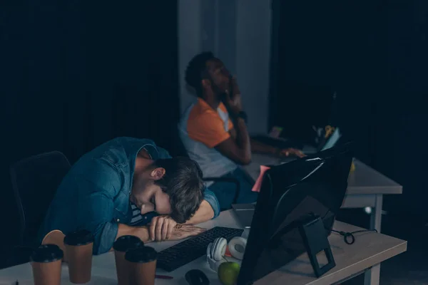 Programmatore esausto dormire sul posto di lavoro mentre collega africano che lavora di notte in ufficio — Foto stock