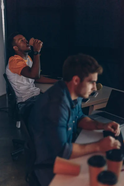 Selective focus of african american programmer drinking coffee to go while working near multicultural colleague at night in office — Stock Photo