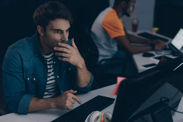 Selective focus of shocked programmer working at night in office near multicultural friend — Stock Photo