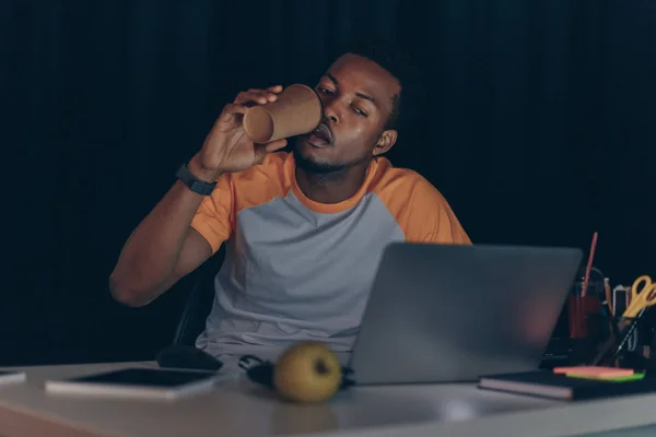 Young african american programmer drinking coffee to go while working at night in office — Stock Photo