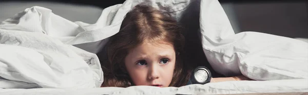 Scared child hiding under blanket with flashlight isolated on black — Stock Photo