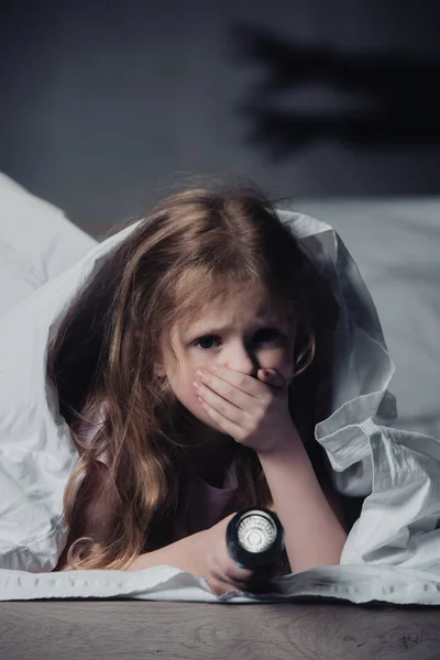 Налякана дитина тримає ліхтарик, ховаючись під ковдрою на чорному тлі з тінями — стокове фото