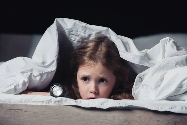 Bambino spaventato nascosto sotto coperta e tenendo torcia isolata su nero — Foto stock