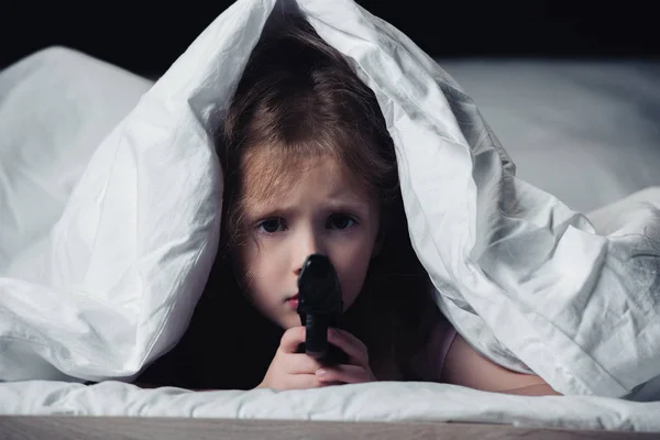 Enfant effrayé tenant pistolet et regardant la caméra tout en se cachant sous une couverture isolée sur noir — Photo de stock
