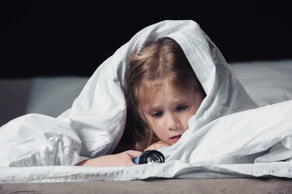 Scared child holding flashlight while hiding under white blanket isolated on black — Stock Photo