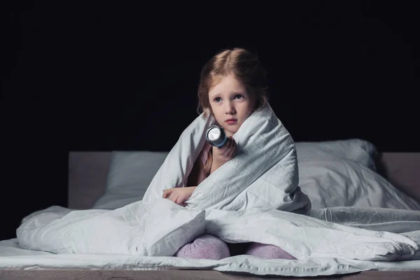 Bambino spaventato seduto sotto coperta e tenendo torcia isolata su nero — Foto stock