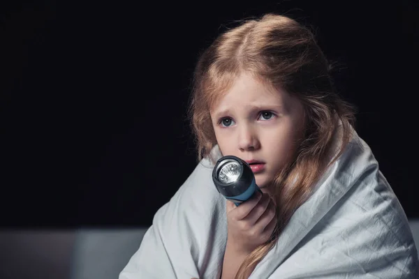 Scared kid sitting under blanket and holding flashlight isolated on black — Stock Photo