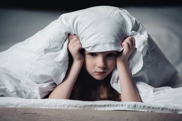 Criança assustada se escondendo sob cobertor e olhando para a câmera isolada no preto — Fotografia de Stock