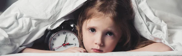Panoramic shot of scared kid hiding under blanket with alarm clock — Stock Photo