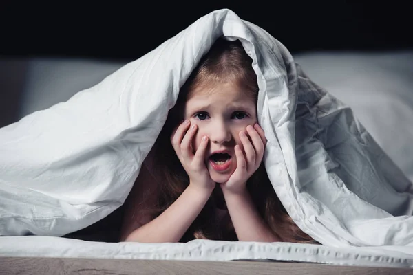 Niño asustado gritando y agarrado de la mano mientras se esconde bajo una manta aislada en negro - foto de stock