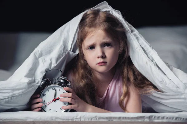 Испуганный ребенок лежит под одеялом с будильником и смотрит на камеру, изолированную на черном — стоковое фото