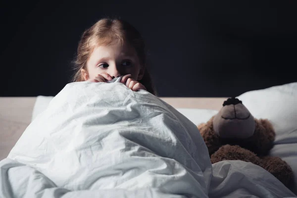 Испуганный ребенок смотрит в сторону, сидя под одеялом рядом с плюшевым мишкой изолированы на черный — стоковое фото