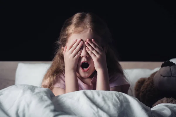Bambino spaventato urlando e coprendo gli occhi con le mani mentre seduto su biancheria da letto vicino orsacchiotto isolato su nero — Foto stock